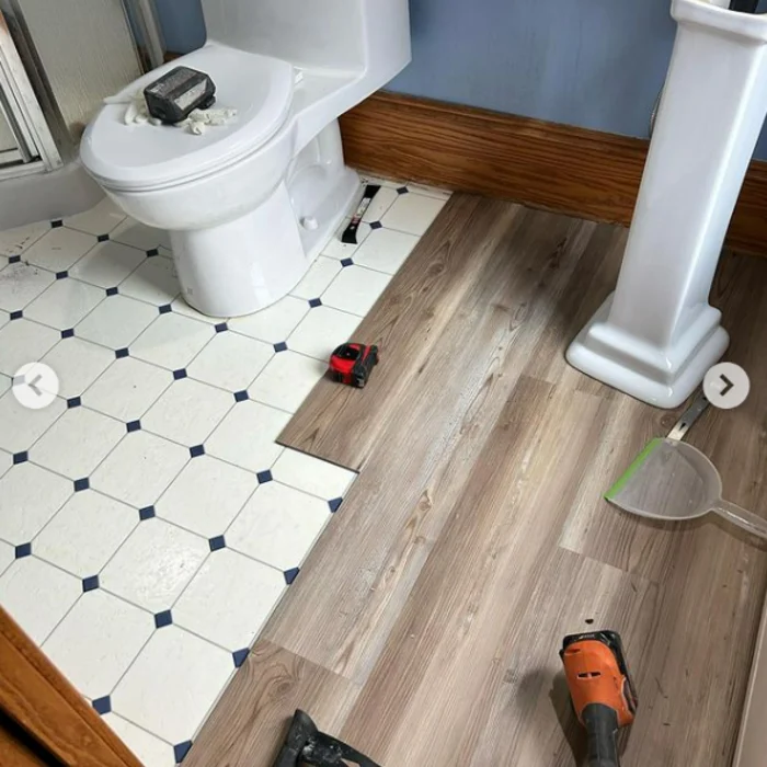 Wood-look flooring partially fitted beside the toilet and pedestal sink, with the old patterned floor still exposed.