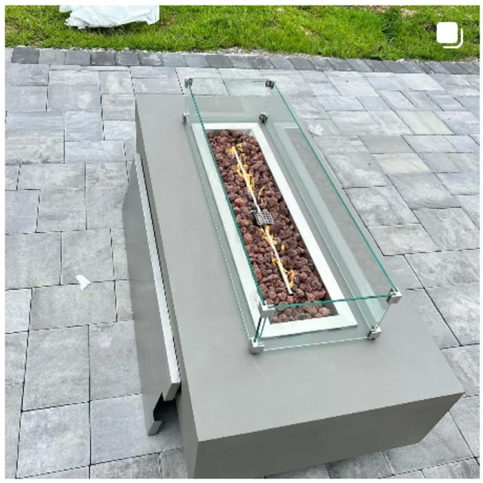 Rectangular outdoor fire table with lava rock and a clear glass wind guard on a paved patio.
